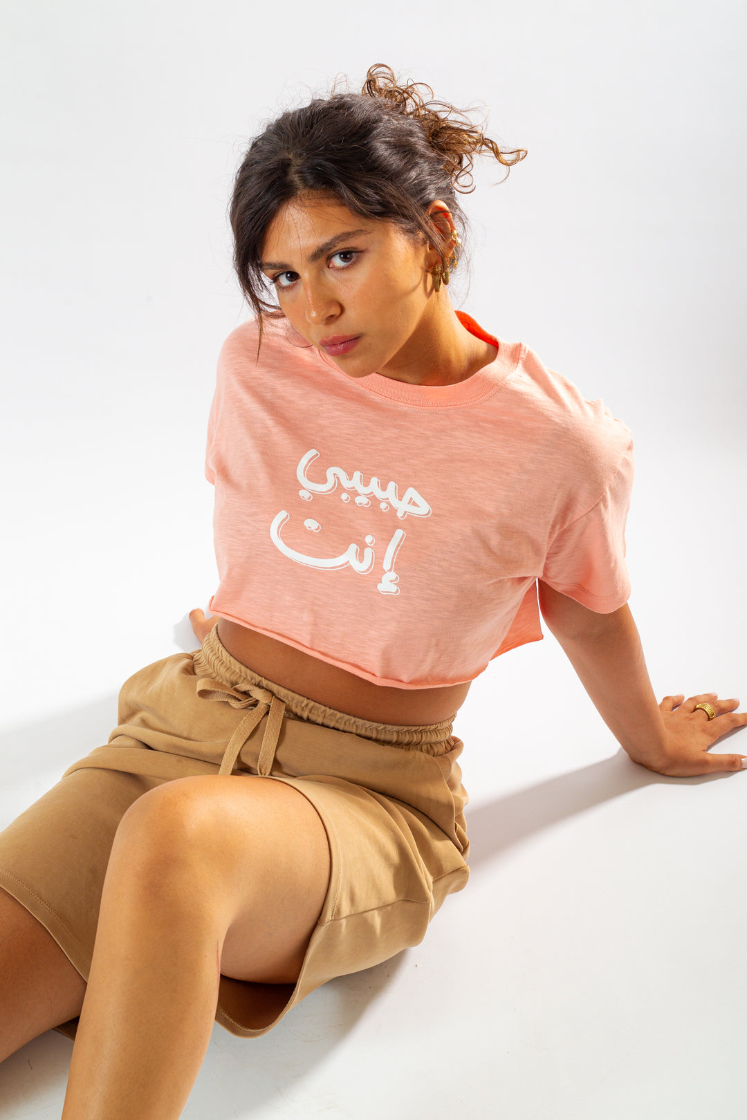 Young adult female wearing an Oversized peach Crop Top with Habibi Enta written in Arabic حبيبي إنت and printed in white silkscreen in the center of the top along with beige shorts.