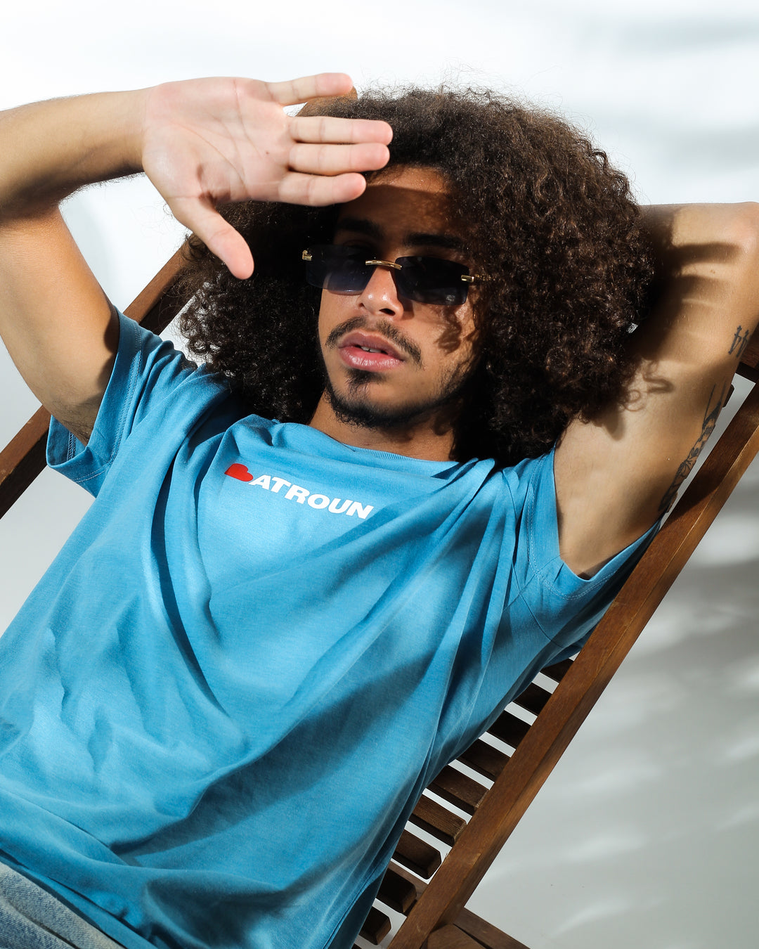Young adult male wearing blue T-shirt with Batroun written in white and printed in silkscreen and the B is replaced with a red heart. He is laying on a wood chair and wearing sunglasses.