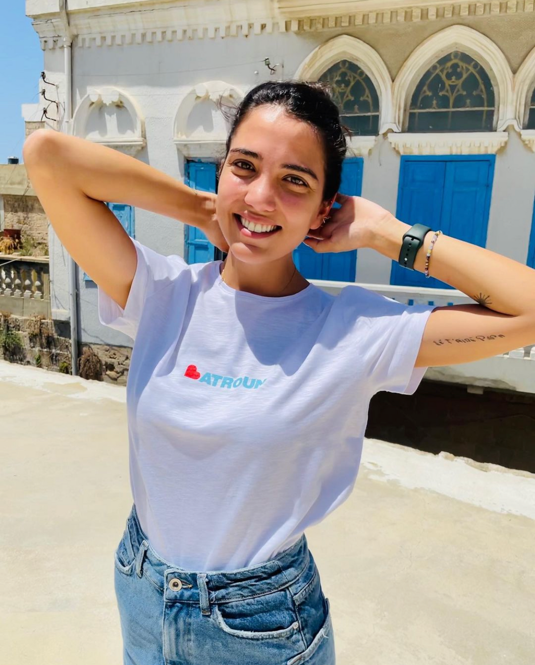 Young adult female wearing white T-shirt with Batroun written in white and printed in silkscreen and the B is replaced with a red heart along with jeans.
