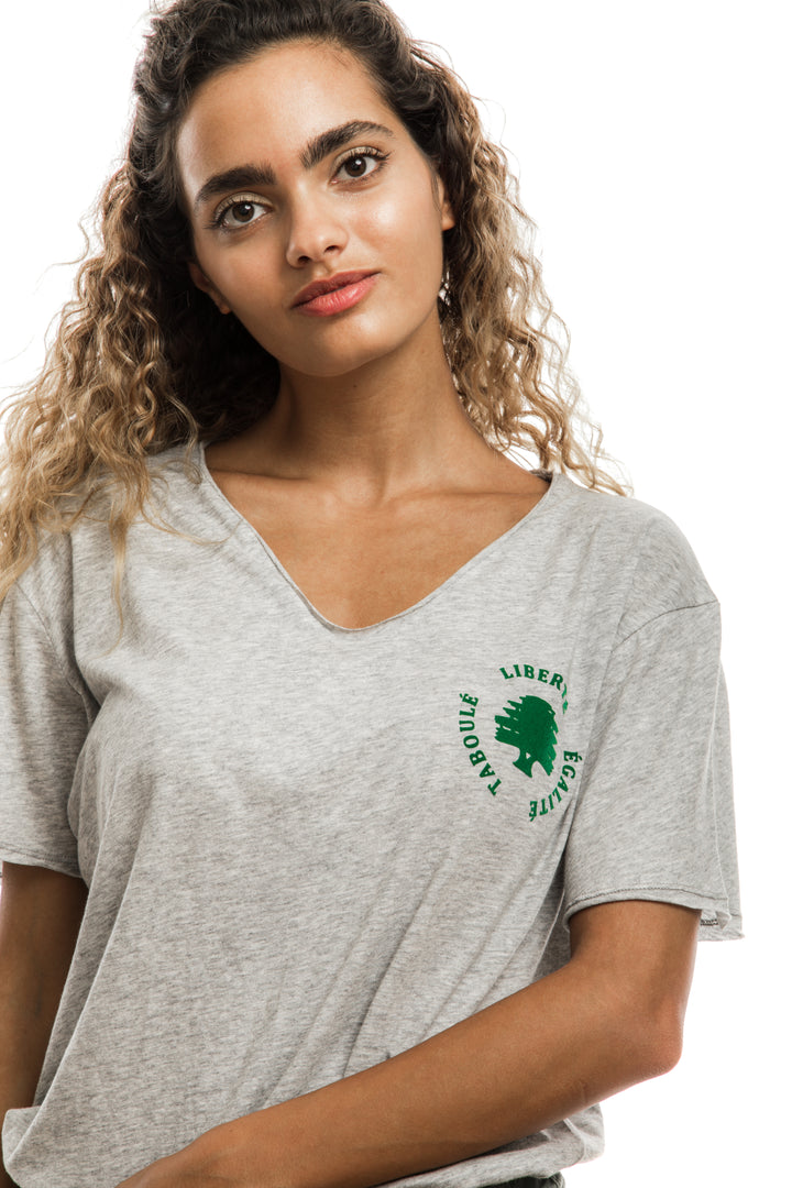 Young adult female wearing grey T-shirt with Liberté Égalité Taboulé written in French and printed in green velvet on the side of the T-shirt.
