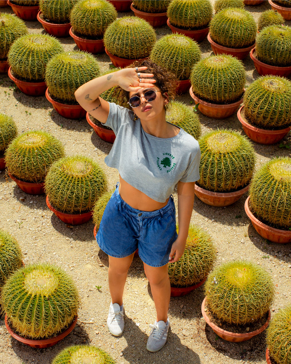 Young adult female wearing grey Crop Top with Liberté Egalité Taboulé written in French and printed in green velvet on the side of the Crop Top along with white sneakers, jeans short and sunglasses. Standing between cactuses in pots.