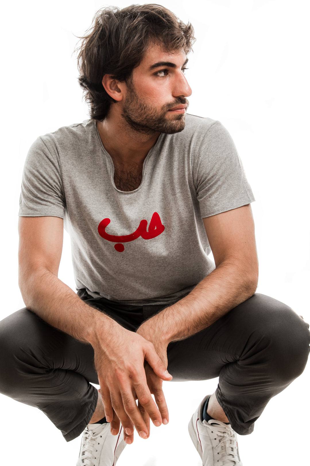 Young adult male wearing grey T-shirt with Hobb written in Arabic حب and printed in red velvet in the center of the T-shirt along with black pants and white sneakers.
