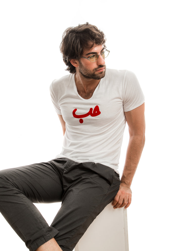 Young adult male wearing white T-shirt with Hobb written in Arabic حب and printed in red velvet in the center of the T-shirt along with black pants and glasses.