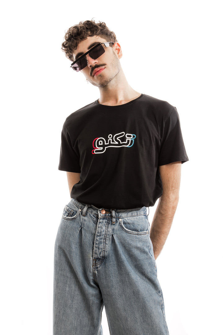 Young adult male wearing black T-shirt with Techno written in Arabic تكنو and printed in blue, red and white silkscreen in the center of the t-shirt along with blue jeans and sunglasses.
