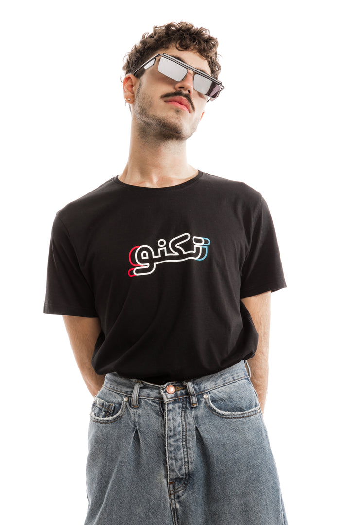 Young adult male wearing black T-shirt with Techno written in Arabic تكنو and printed in blue, red and white silkscreen in the center of the t-shirt along with blue jeans and sunglasses.