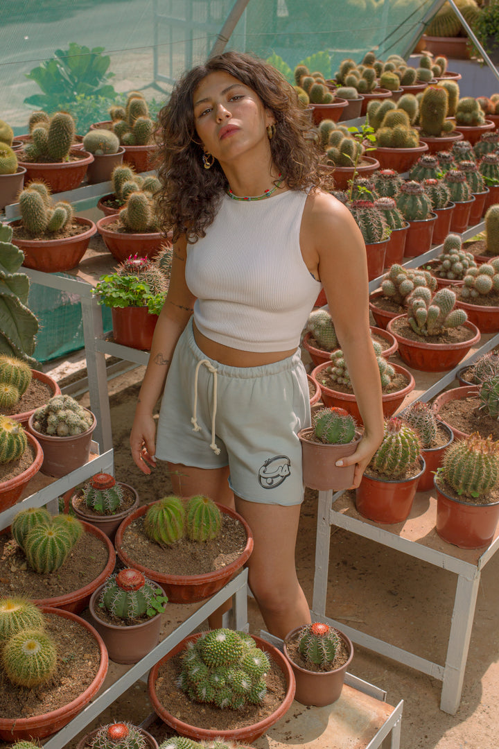Young adult female wearing grey Flared Short with Kell el Hobb written in Arabic كل الحب and printed in black silkscreen on the side of the short along with a white sleeveless top. Standing between cactus pots and holding one in her left hand.
