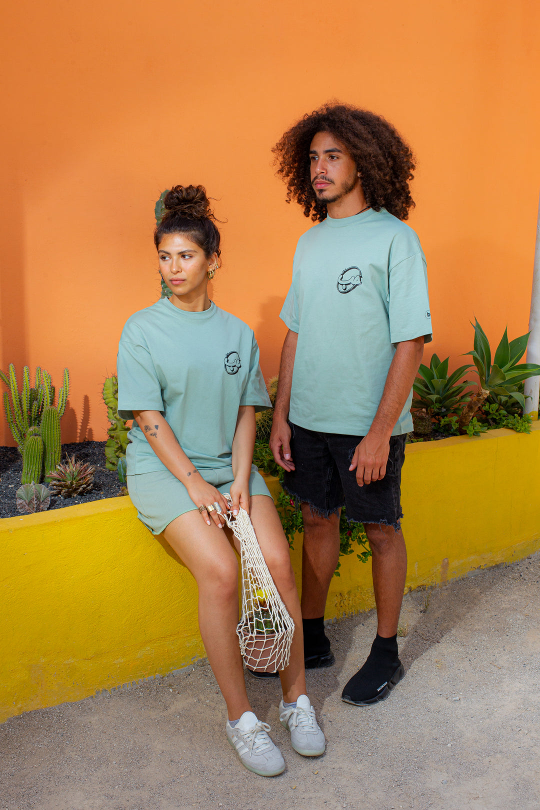 Young adult male and female wearing an Oversized mint T-shirt with Kell el Hobb written in Arabic كل الحب and printed in black silkscreen. They are both in front of an orange wall. She is wearing the T-shirt along with a mint short, grey sneakers and holding a pot with a cactus in a net. He is standing next to her wearing the T-shirt with black jeans short and black running shoes.