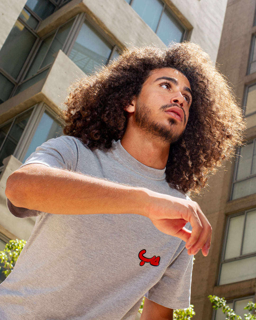 Young adult male wearing an Oversized grey T-shirt with Hobb written in Arabic حب and printed in red silkscreen on the side of the shirt. Standing in front of a modern building.