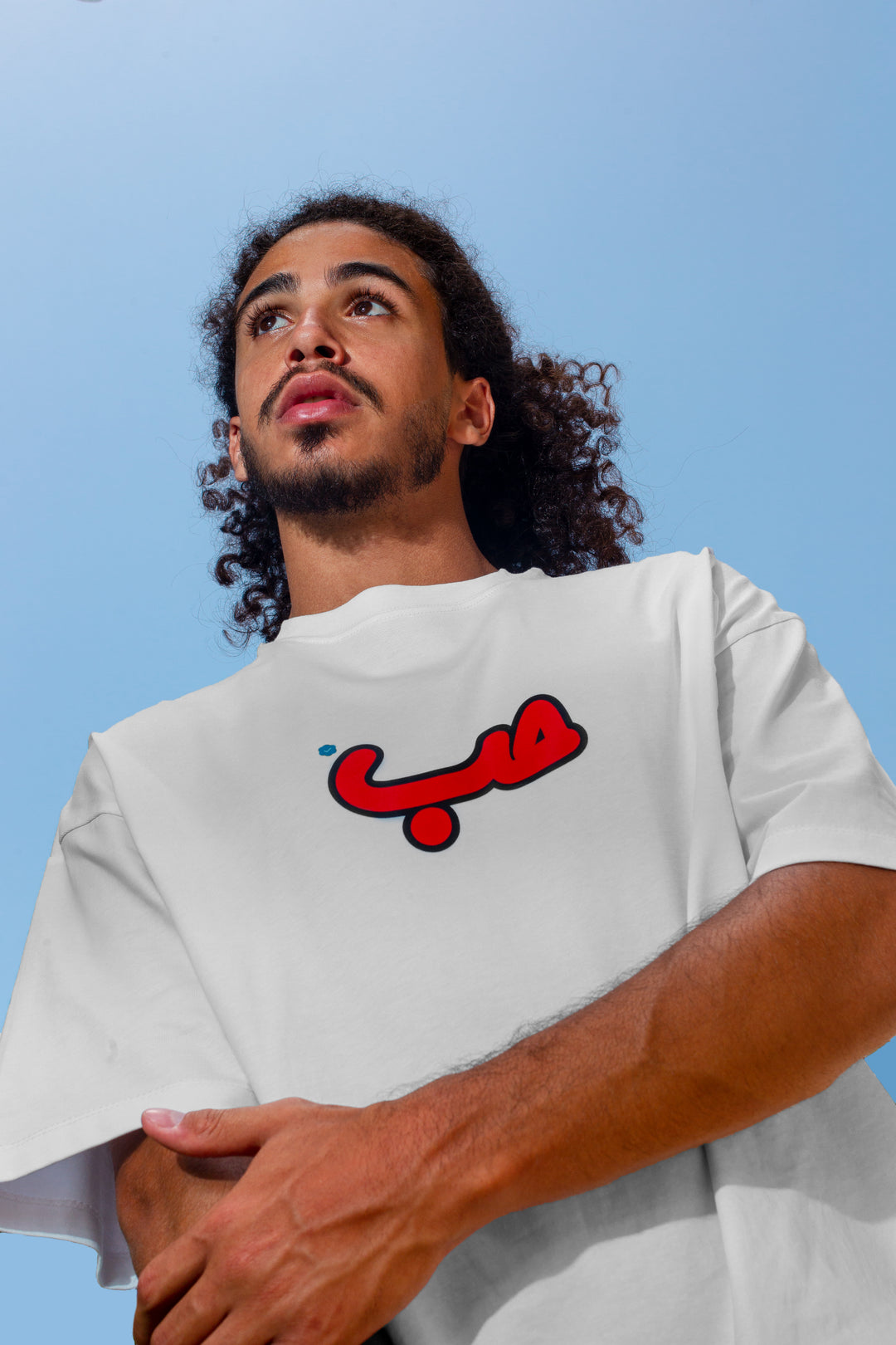 Young adult male wearing an Oversized white T-shirt with Verified Hobb written in Arabic حب and printed in red silkscreen in the center of the shirt.
