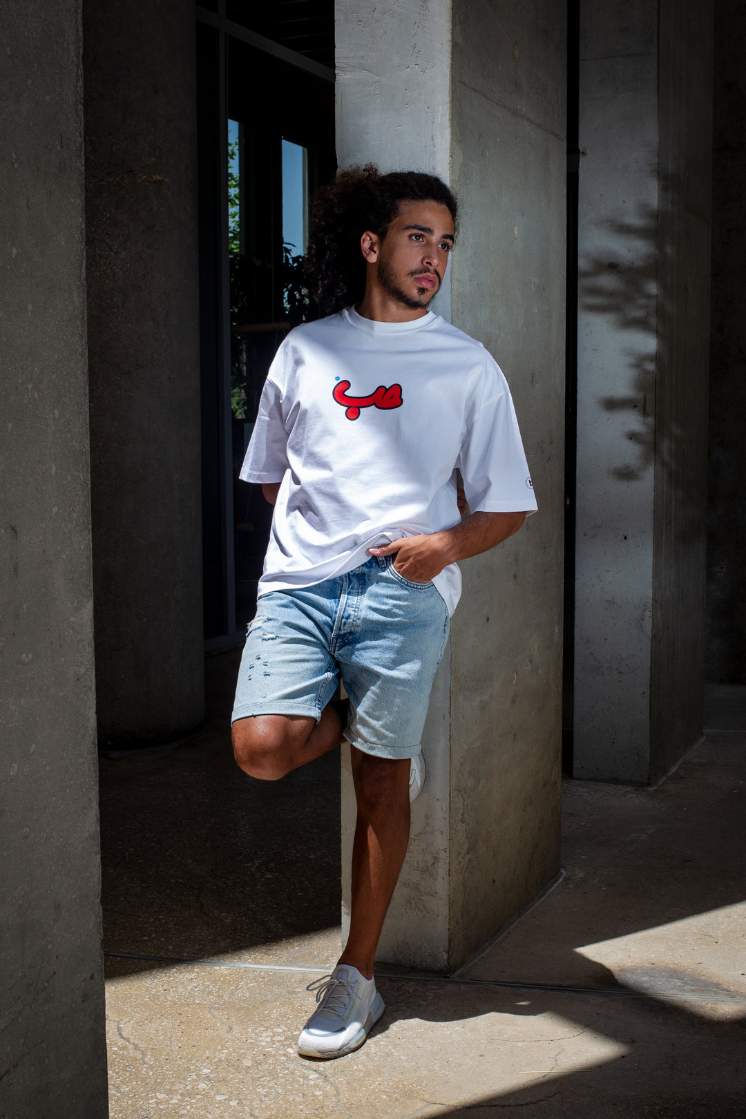 Young adult male wearing an Oversized white T-shirt with Verified Hobb written in Arabic حب and printed in red silkscreen in the center of the shirt along with jeans shorts and white sneakers.