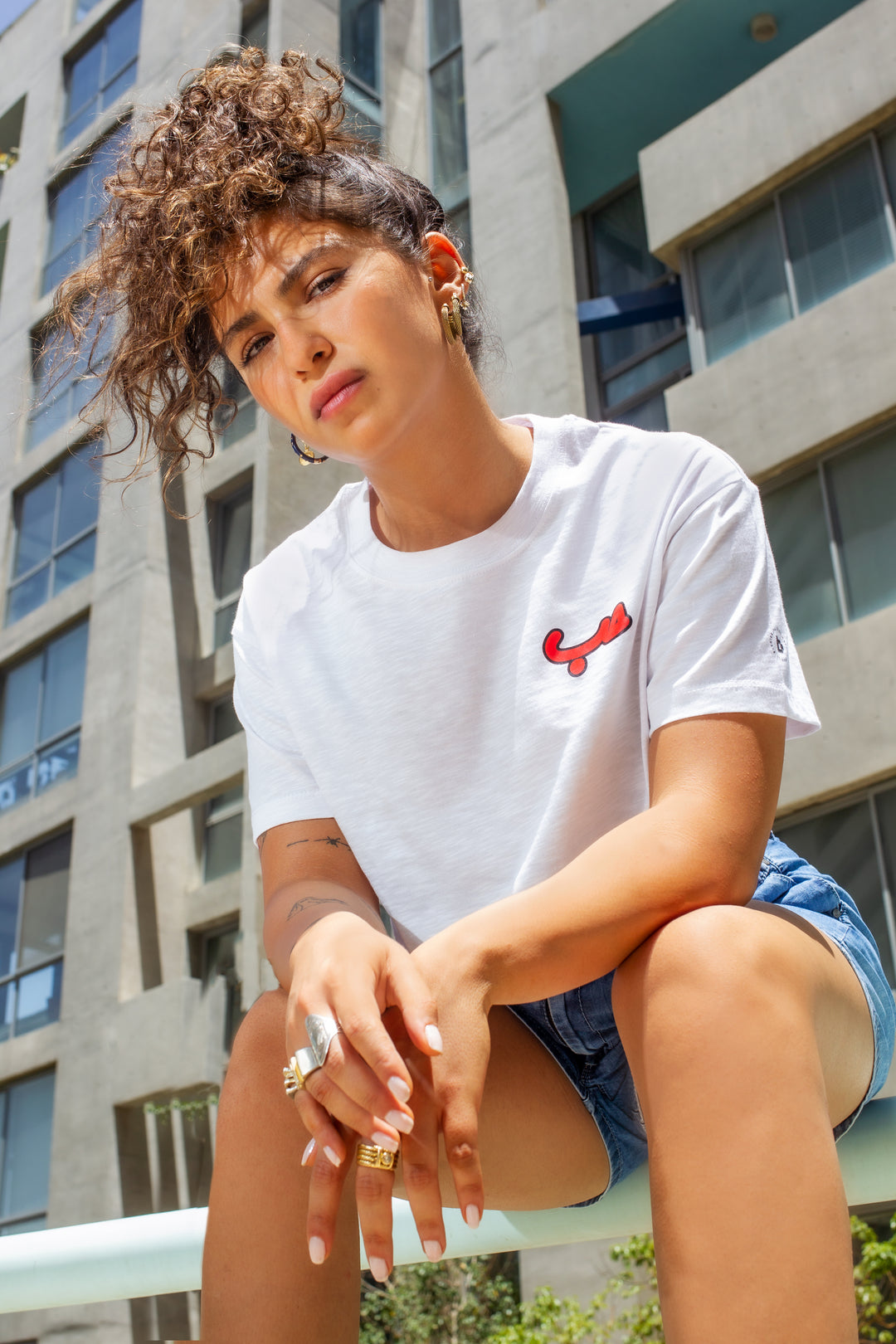 Young adult female wearing an Oversized white Crop Top with Hobb written in Arabic حب and printed in red silkscreen in the center of the shirt along with jeans shorts. Sitting in front of a modern building.