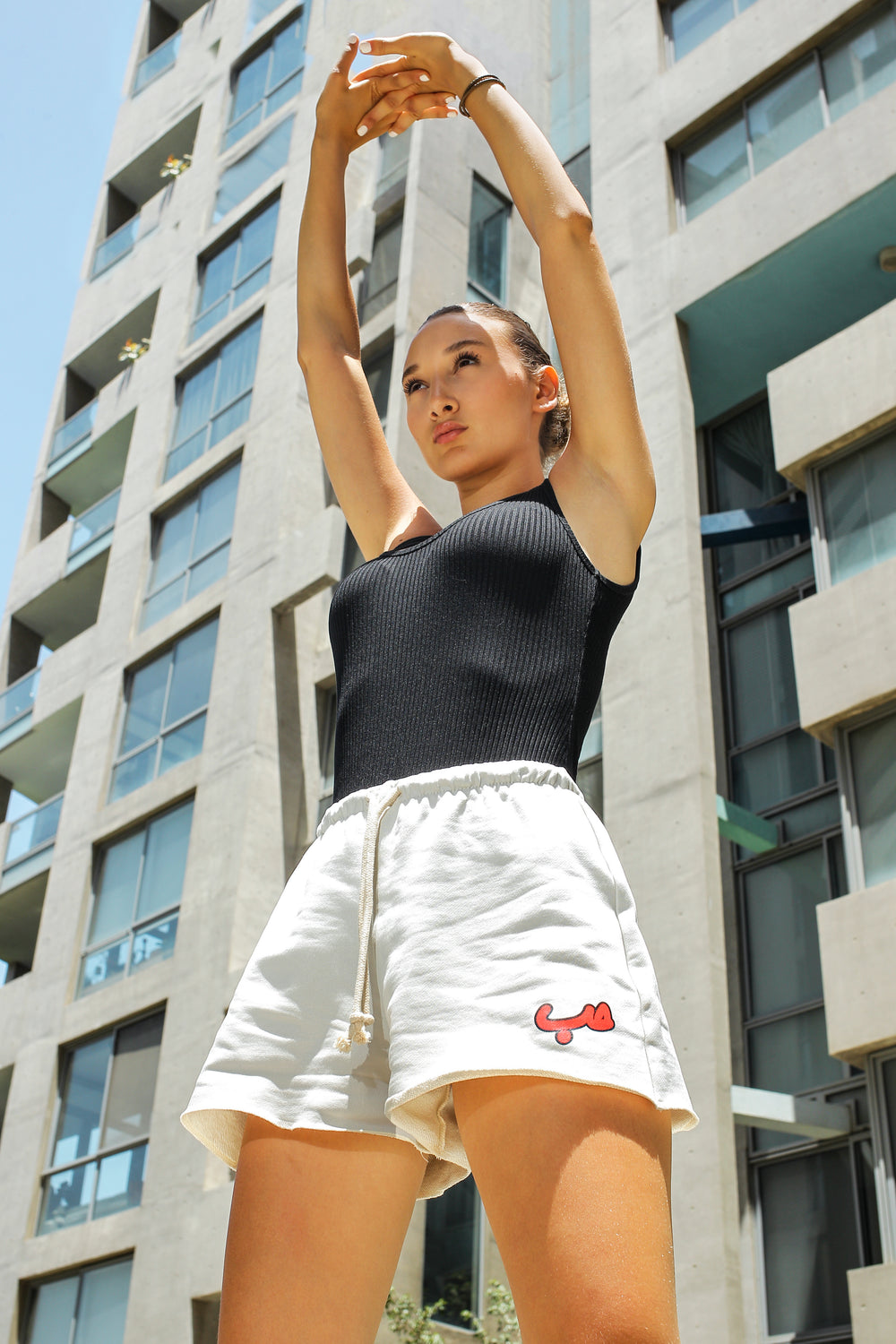 Young adult female wearing off white Flared Short with Hobb written in Arabic حب and printed in red silkscreen on the side of the short along with a black sleeveless top. Standing in front of a modern building.