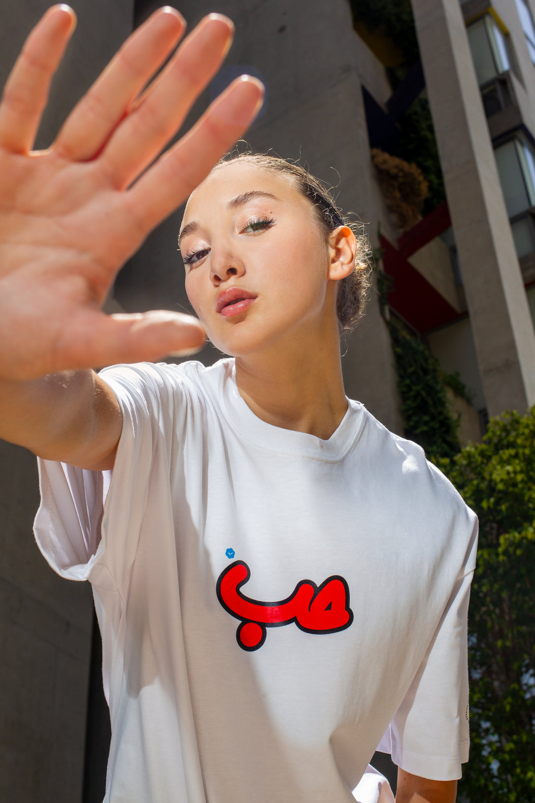 Young adult female wearing an Oversized white T-shirt with Verified Hobb written in Arabic حب and printed in red silkscreen in the center of the shirt. Standing in front of a modern building.