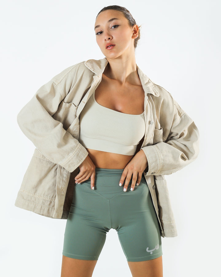 Young adult female wearing bay leaf Biker Short with Hobb written in Arabic حب and printed in reflective silver on the side of the short along with an off white sleeveless top and a beige jacket.