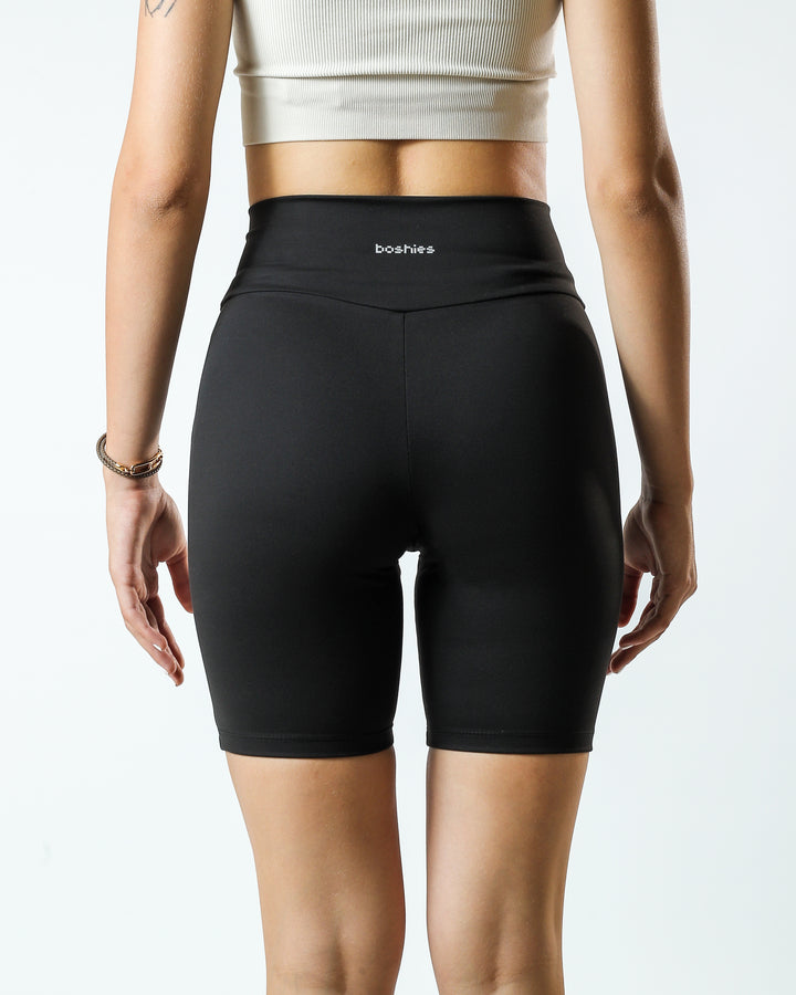 Close up on a young adult female, seen from the back, wearing black Biker Short with Boshies written on the top center of the short and printed in reflective silver along with an off white top.