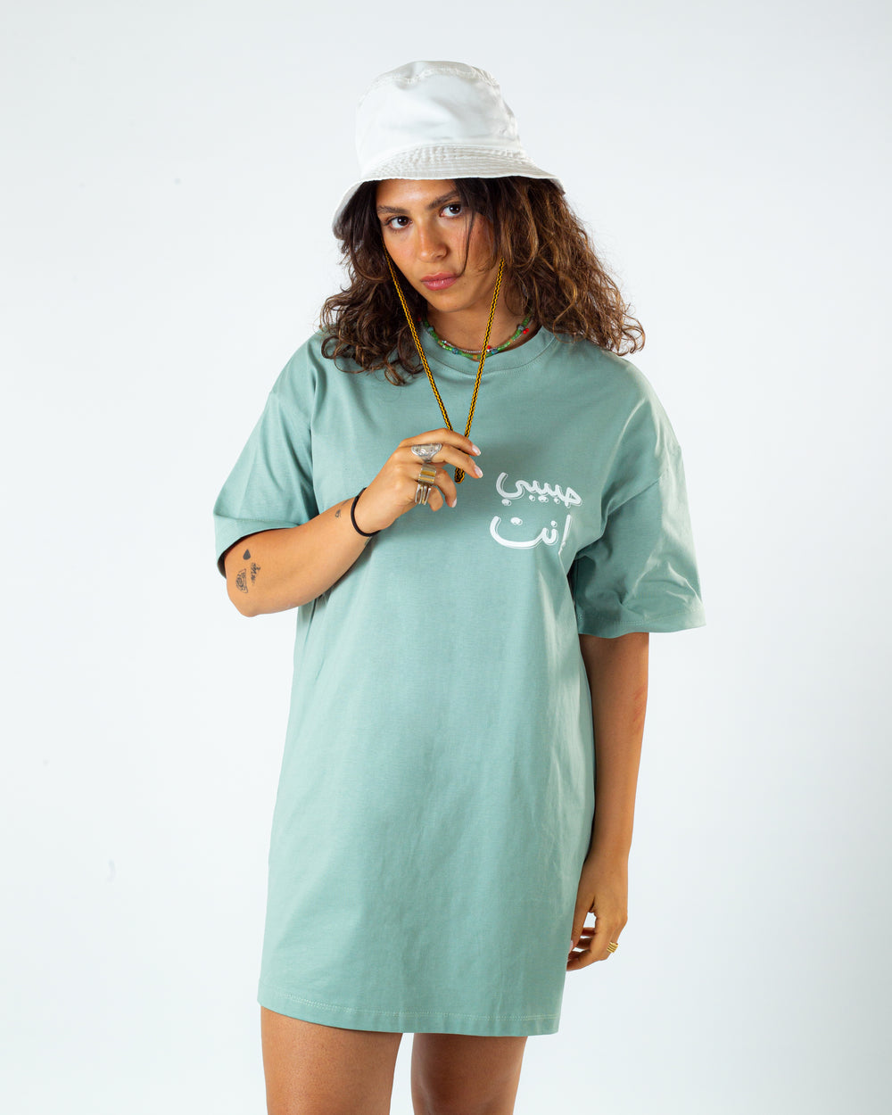 Young adult female wearing an Oversized mint T-dress with Habibi Enta written in arabic حبيبي إنت and printed in white silkscreen on the side of the T-Dress along with a white hat on her head.