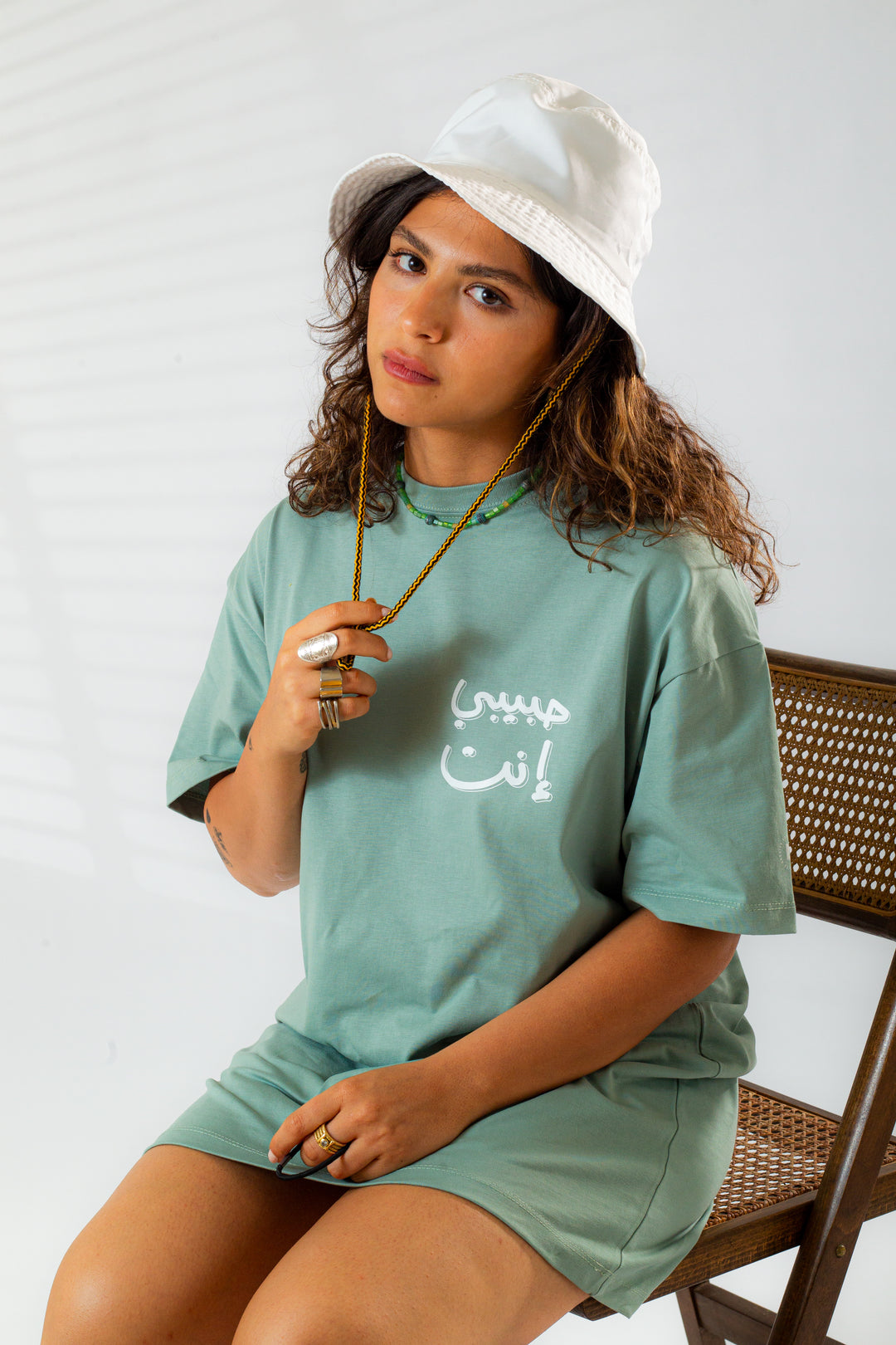 Young adult female wearing an Oversized mint T-dress with Habibi Enta written in Arabic حبيبي إنت and printed in white silkscreen on the side of the T-Dress along with a white hat on her head. Sitting on a wood chair.