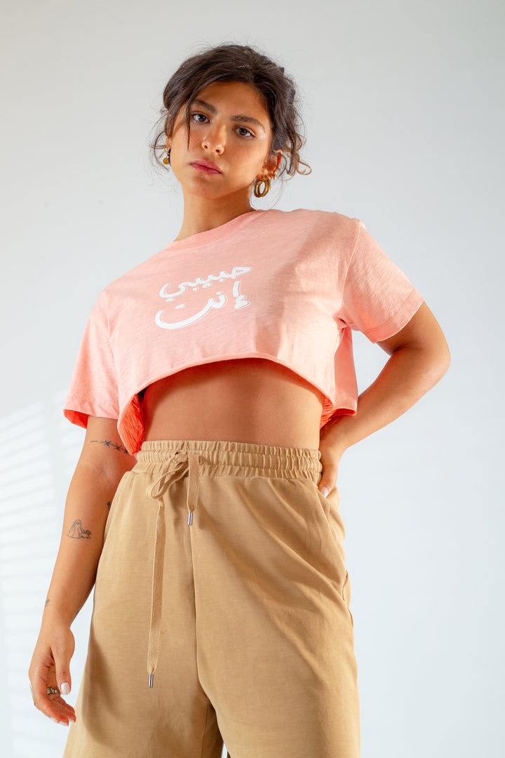Young adult female wearing an Oversized peach Crop Top with Habibi Enta written in Arabic حبيبي إنت and printed in white silkscreen in the center of the top along with beige shorts.