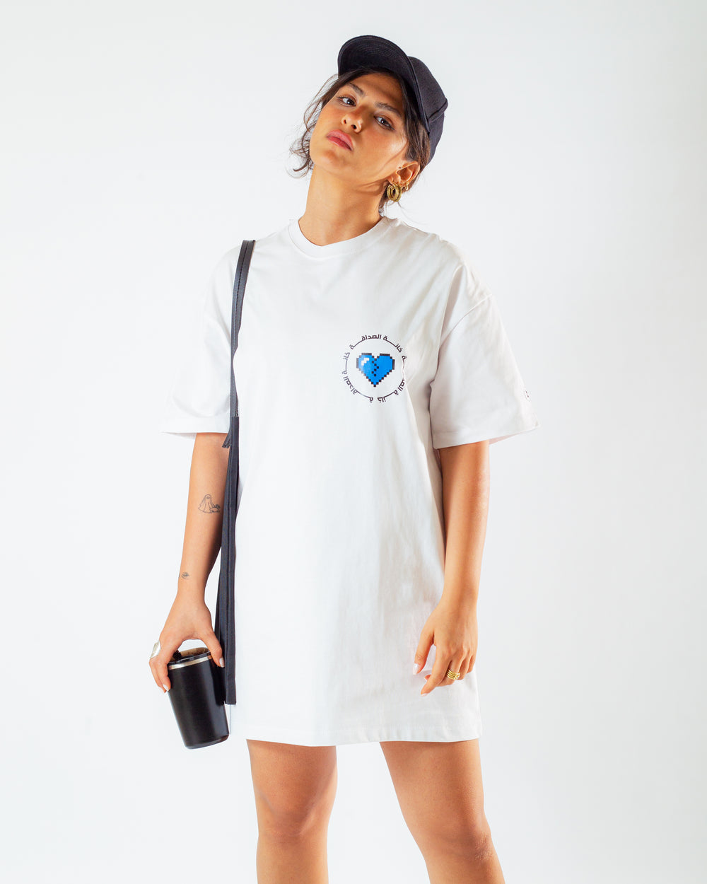 Young adult female wearing an Oversized white T-Dress with Khanat Al Sadaka written in arabic خانة الصداقة and printed in blue and black silkscreen on the side of the dress along with black cap on her head, a black tote bag and a black coffee cup.