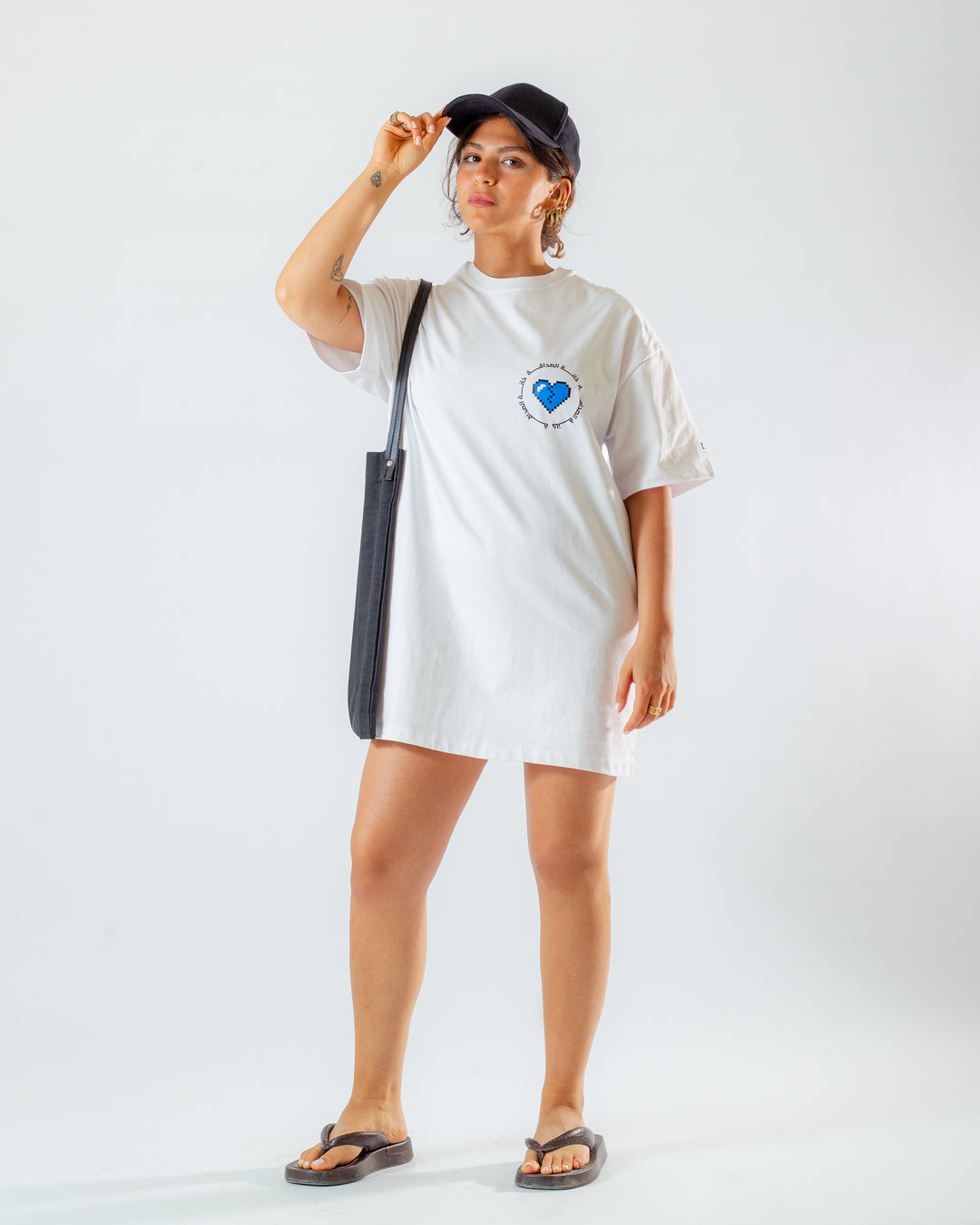 Young adult female wearing an Oversized white T-Dress with Khanat Al Sadaka written in arabic خانة الصداقة and printed in blue and black silkscreen on the side of the dress along with a black cap on her head and a black tote bag.