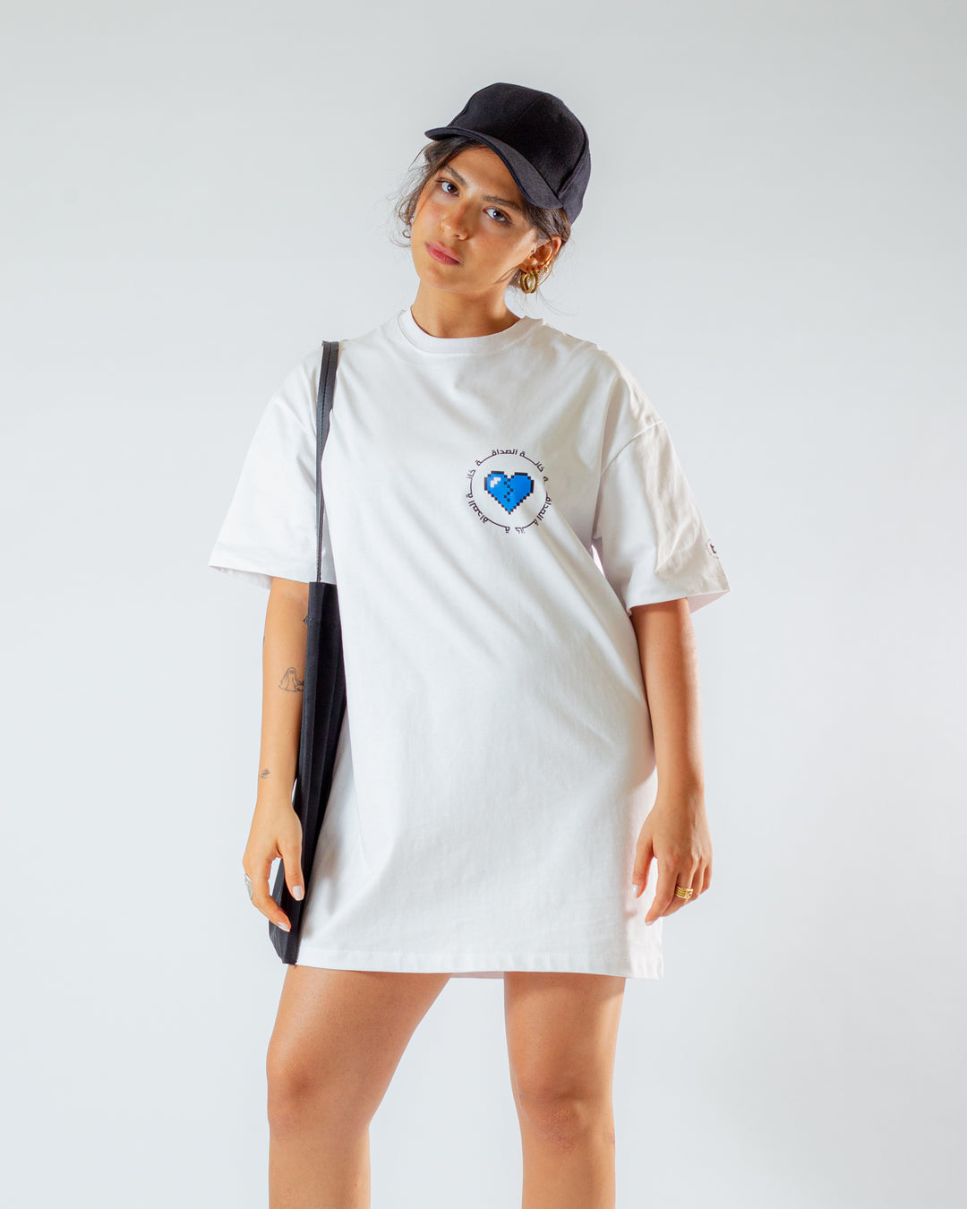 Young adult female wearing an Oversized white T-Dress with Khanat Al Sadaka written in Arabic خانة الصداقة and printed in blue and black silkscreen on the side of the dress along with a black cap on her head and a black tote bag.