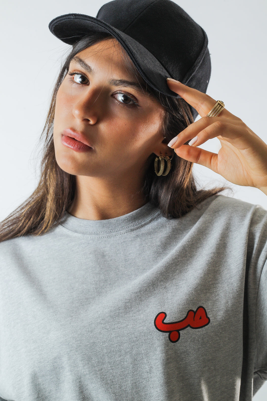 Young adult female wearing an Oversized grey T-Dress with Hobb written in Arabic حب and printed in red silkscreen on the side of the dress along with a black cap on her head.