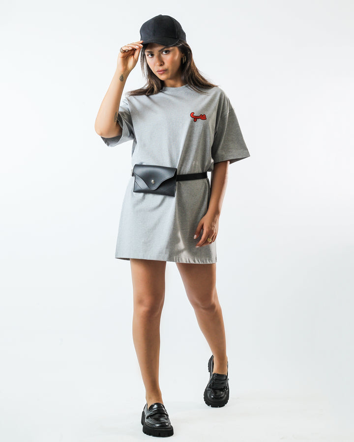 Young adult female wearing an Oversized grey T-Dress with Hobb written in Arabic حب and printed in red silkscreen on the side of the dress along with a black cap, a black belt bag and black shoes.