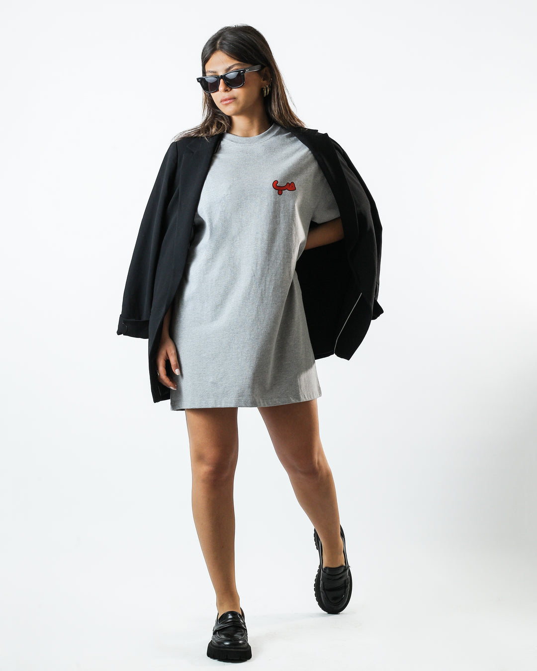 Young adult female wearing an Oversized grey T-Dress with Hobb written in Arabic حب and printed in red silkscreen on the side of the dress along with a black jacket, black shoes and sunglasses.