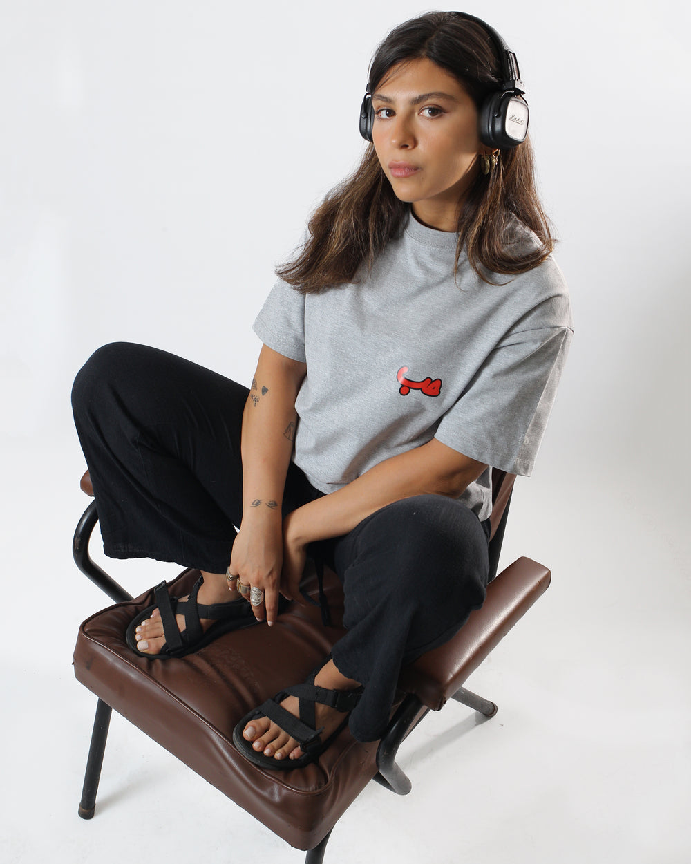 Young adult female wearing an Oversized grey T-shirt with Hobb written in Arabic حب and printed in red silkscreen on the side of the shirt along with black pants, black sandals and black headphone. Sitting on a brown leather chair.