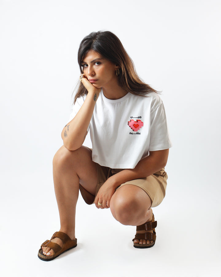Young adult female wearing white Crop Top with Albi 3a Albak written in Arabic قلبي ع قلبك and printed pink in silkscreen along with long beige shorts and brown sandals.
