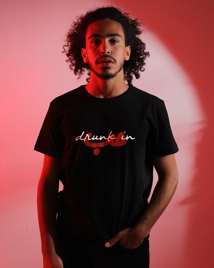 Young adult male wearing black T-Shirt with Drunk in Hobb written in English and Arabic حب and printed in white and red silkscreen along with black jeans.