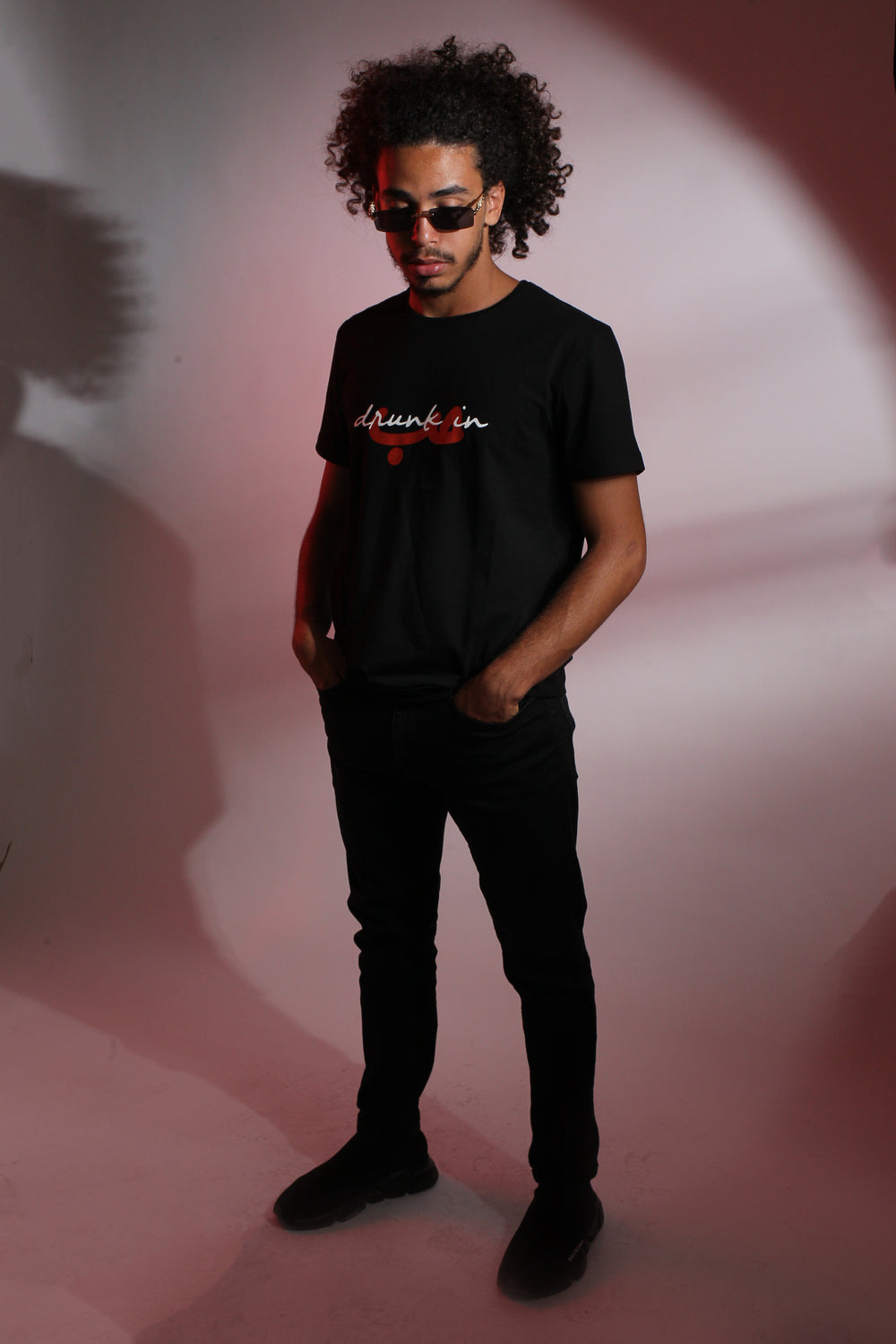 Young adult male wearing black T-Shirt with Drunk in Hobb written in English and Arabic حب and printed in white and red silkscreen along with black jeans, black shoes and sunglasses.