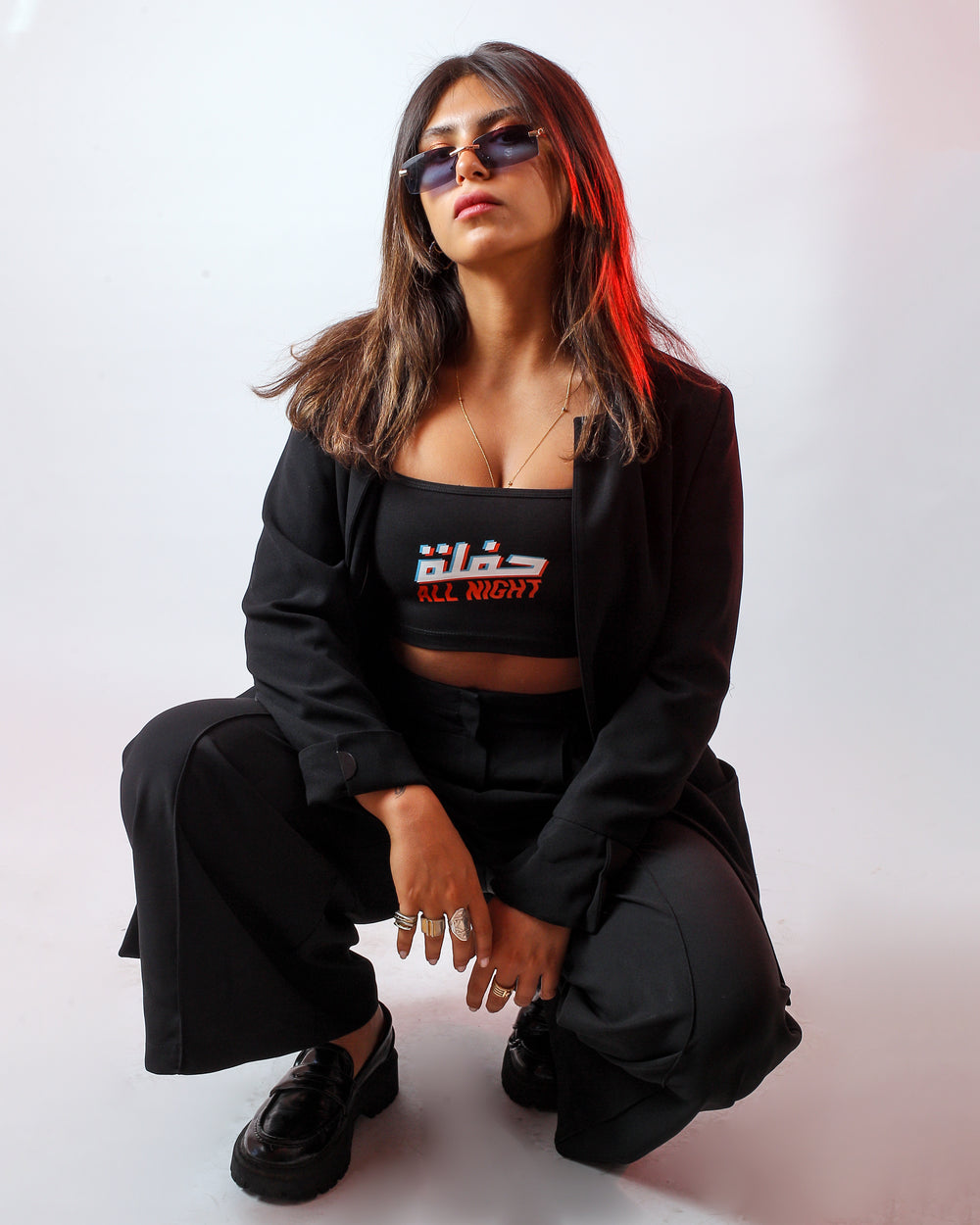 Young adult female wearing black Bralette with Hafle all Night written in Arabic and English حفلة All Night and printed in glitch silkscreen in the center of the bralette along with black trousers, black shoes, a black jacket and sunglasses.