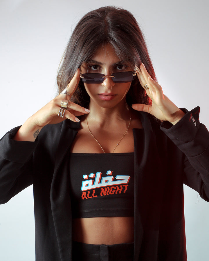 Young adult female wearing black Bralette with Hafle all Night written in Arabic and English حفلة All Night and printed in glitch silkscreen in the center of the bralette along with a black jacket and sunglasses.
