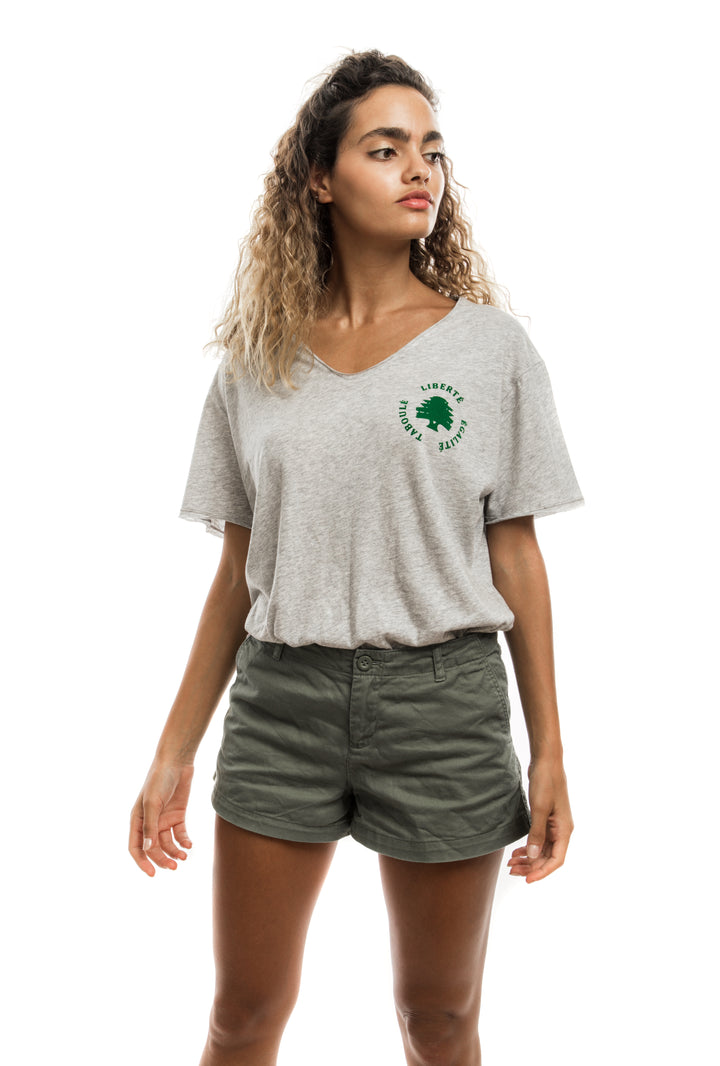 Young adult female wearing grey T-shirt with Liberté Égalité Taboulé written in French and printed in green velvet on the side of the T-shirt along with olive green shorts.