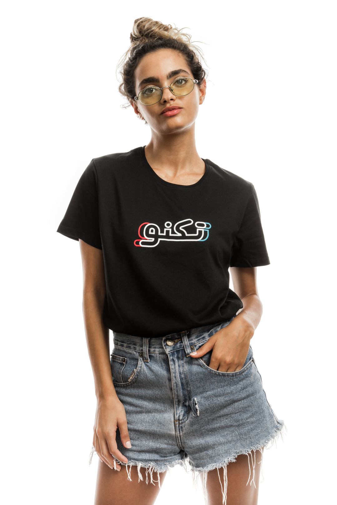 Young adult female wearing black T-shirt with Techno written in Arabic تكنو and printed in blue, red and white silkscreen in the center of the t-shirt along with blue jeans shorts and glasses.
