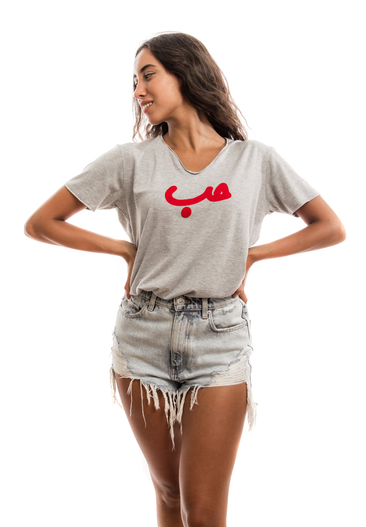young adult female wearing grey t-shirt with Hobb written in arabic حب and in red velvet in the center of the t-shirt along with light blue jeans shorts.