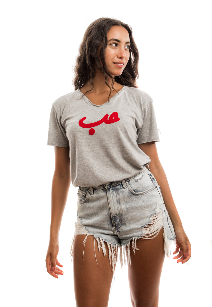 Young adult female wearing grey T-shirt with Hobb written in Arabic حب and printed in red velvet in the center of the T-shirt along with light blue jeans shorts.