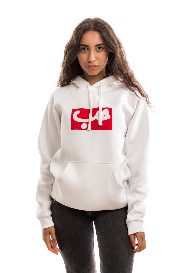 Young adult female wearing white Hoodie with Hobb written in Arabic حب in white and printed in a red velvet frame in the center of the Hoodie along with black jeans.
